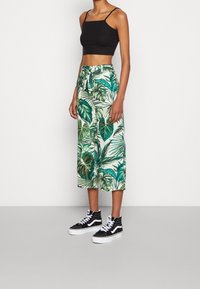Débardeur noir raccourci associé à un pantalon large imprimé tropical vert avec cordon de serrage, porté avec des baskets montantes noires.