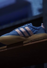 Blue sneaker with white stripes and beige sole resting on a wooden surface against a dark blurred background.
