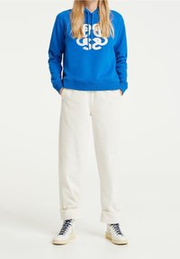 Blue hoodie with white graphic logo, paired with cream corduroy pants. Model wears white sneakers with navy laces. Casual streetwear style.