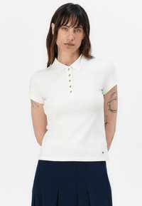 Bílá polo košile s krátkými rukávy a límečkem, s zlatými knoflíky a malým logem na lemu. Hladká textura látky.