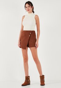 Haut blanc à col montant côtelé associé à une mini-jupe en suède marron avec un design enroulé diagonal et deux grands boutons sur le devant. Bottes chelsea marron.