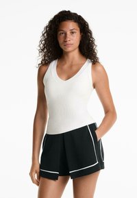 Débardeur blanc à côtes avec un col en V, associé à un short noir avec des bordures blanches et des poches, mettant en avant une coupe décontractée et un design plissé.