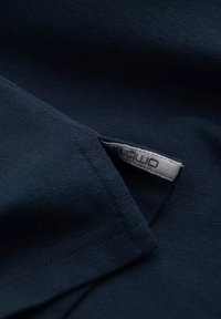 Tejido texturizado de color azul marino con un borde doblado, que presenta una pequeña etiqueta blanca cosida, mostrando el nombre de la marca.