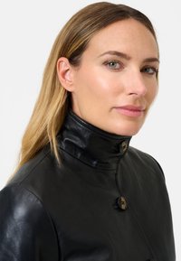 Zwarte leren jas met een hoge kraag, voorzien van een gladde textuur, knopen aan de voorkant en een getailleerd ontwerp. Minimalistische details door het geheel.