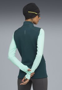 Gilet aderente con zip di colore verde scuro e maniche lunghe verde menta. Tessuto morbido e cuciture a contrasto, con colletto alto e aperture per il pollice.