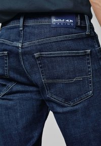 Tamsiai mėlynos džinsinės kelnės su tiesiu kirpimu, turinčios dvi užpakalines kišenes ir ženklo odinę etikete juosmens srityje.