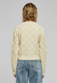 Crème-kleurige gebreide trui met een gestructureerd dambordpatroon, voorzien van lange mouwen en een geribbelde tailleband, gecombineerd met lichtblauwe jeans.