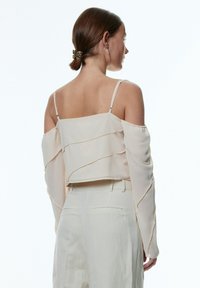 Helle cremefarbene Bluse mit schulterfreier Form, geschichteten Rüschen und langen Ärmeln. Hergestellt aus leichtem Stoff und verfügt über feine Nähte entlang der Kanten.