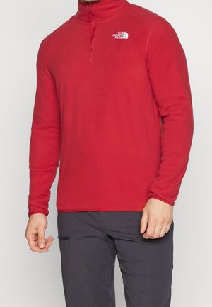 Mężczyzna w czerwonej polarowej bluzie z zamkiem do połowy i logo The North Face na piersi oraz w ciemnoszarych spodniach.