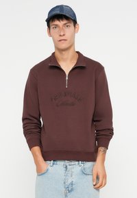 Bordowy dres z pół zamkiem, z haftowanym napisem "ORIGINALS" na klatce piersiowej. Materiał bawełniany; luźny krój, długie rękawy, teksturowana tkanina.