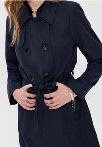 Marinblå trenchcoat med dubbelknäppning, stora svarta knappar och bälte i midjan. Slät tyg med skräddarsydd passform.