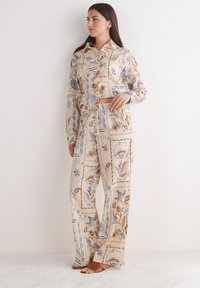 Outfit a due pezzi in tessuto beige chiaro presenta un design a motivo con accenti floreali in blu e marrone. Include una camicia con colletto e pantaloni larghi.