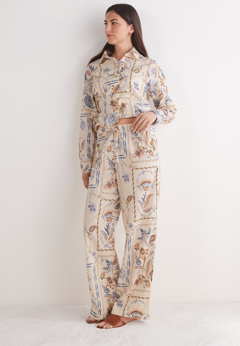 Outfit a due pezzi in tessuto beige chiaro presenta un design a motivo con accenti floreali in blu e marrone. Include una camicia con colletto e pantaloni larghi.