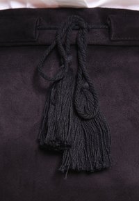 Svart mocka väska med en texturerad yta, med två svarta tofsar fästa vid det övre öppningen. Tofsarna  är täta och fransiga i ändarna.
