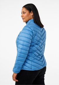 Giacca puffer blu chiaro con colletto alto, caratterizzata da un motivo a zigzag trapuntato e polsini elastici. Indossata con pantaloni neri.