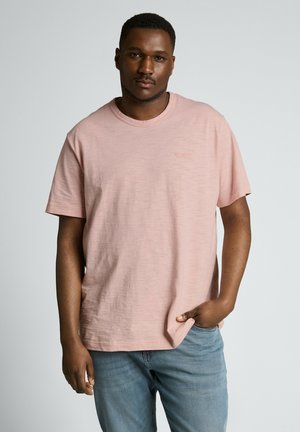 Man die een lichtroze T-shirt met korte mouwen en een blauwe spijkerbroek draagt, staand met één hand in de zak tegen een effen achtergrond.