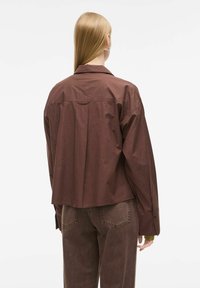 Bruine katoenen shirt met een ontspannen pasvorm, voorzien van een knoopsluiting bij de kraag, brede mouwen en een rugplooidetail, gecombineerd met donkerbruine jeans.