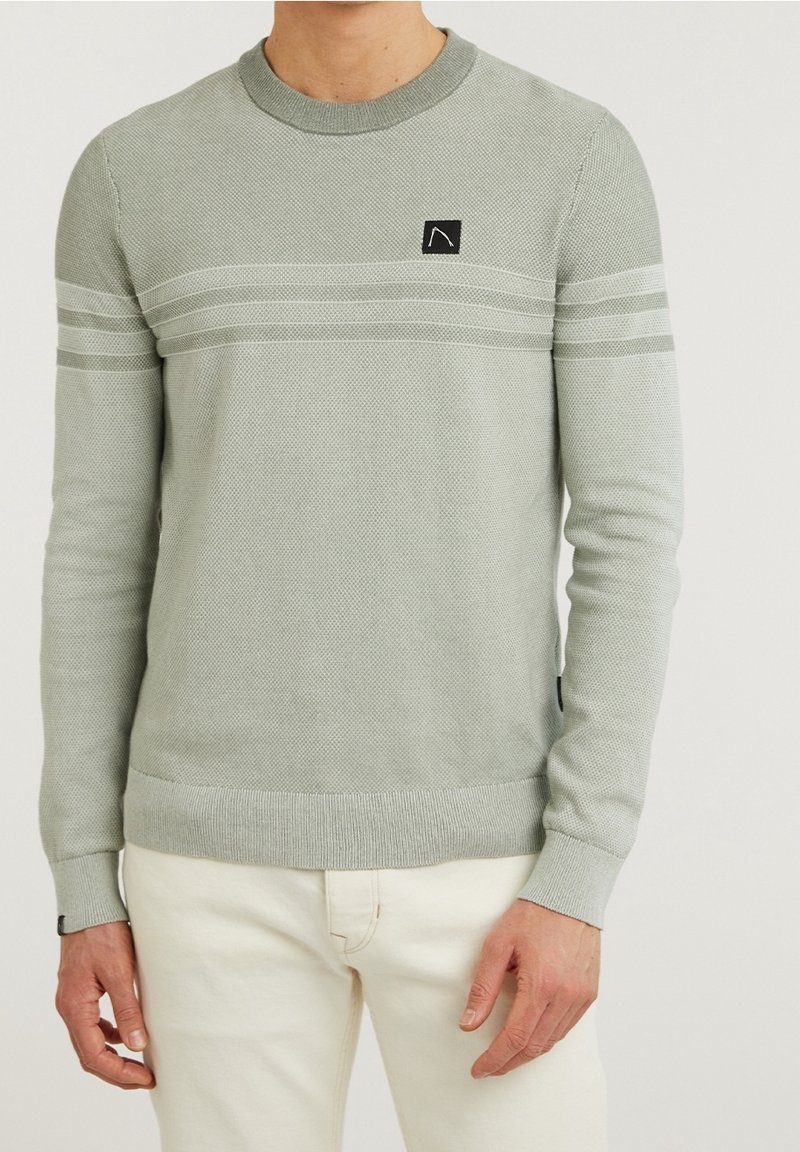 Homme portant un pull à manches longues gris clair texturé avec deux bandes horizontales sur la poitrine et un pantalon beige.