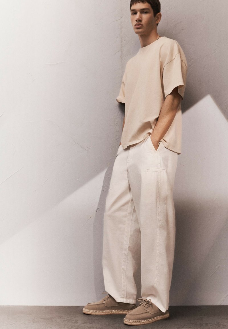 Beige, oversized póló laza fehér nadrággal és világosbarna cipővel párosítva. Egyszerű, letisztult dizájn, laza szabásban és minimális részletezéssel.