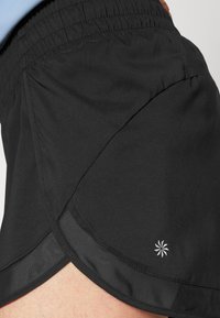 Bermudas atléticas pretas com um material leve, uma cintura elástica e uma bainha curva, apresentando um pequeno logótipo branco na lateral.