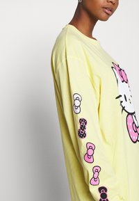 Camiseta de manga larga amarilla hecha de algodón, con un gran gráfico de Hello Kitty en la parte frontal y pequeños lazos a lo largo de las mangas.