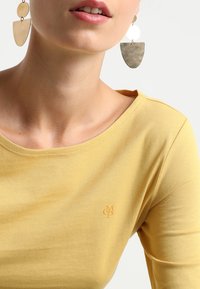 Nainen, joka käyttää suuria geometrisiä kultaketjuja ja keltaista pyöreäkaulaista paitaa, jossa on pieni brodattu logo rinnassa.