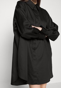 Femme portant une robe chemise noire oversize à manches longues, col large et bras croisés sur fond uni.