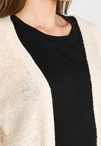 Svart ribbad stickad topp med en öppen, lätt beige stickad kofta med en texturerad mönster och avslappnad passform.