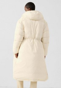 Crèmekleurige donsjas met capuchon, elastische tailleband en lange mouwen; heeft een doorgestikt patroon en een gladde textuur.