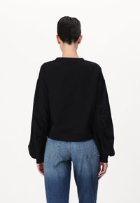 Melna puloverjaketa ar garām piedurknēm, ribotām apakšmalām un sānu burzījuma detaļām, apvienota ar gaiši zilām augstās jostas vietas džinsām.