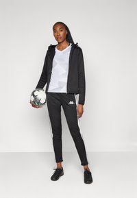 Zwarte zip-up jack over een wit patroon shirt, gecombineerd met zwarte broek met witte zijkanten. Houdt een zwart-wit voetbal vast.