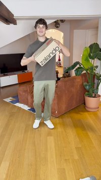 Καφέ χαρτόκουτο με μαύρο κείμενο "zalando", κρατούμενο από ένα άτομο που στέκεται σε ξύλινο δάπεδο σε ένα σαλόνι με καναπέ και φυτά.