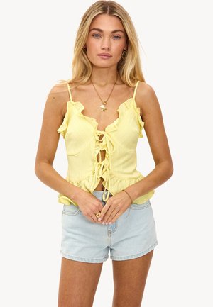 Femme blonde portant un haut sans manches à volants jaune avec un nœud à l'avant et un short en denim bleu clair, debout avec les mains jointes.