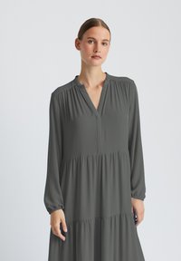 Robe grise à manches longues avec encolure à patte de boutonnage, détails froncés aux épaules, et jupe à volants en matière douce et fluide.