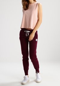 Roze mouwloze crop top gecombineerd met bordeaux joggers met een witte trekkoord en een blauwe logo patch. Witte sneakers maken de outfit compleet.