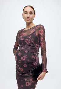 Εγκυμονούσα φόρεμα, διάφανα μακριά μανίκια, σκούρο ύφασμα με ροζ λουλουδάτο μοτίβο, λεπτομέρειες με ρελιού στα πλαϊνά της μέσης, συνδυασμένο με μαύρο τσαντάκι χειρός.