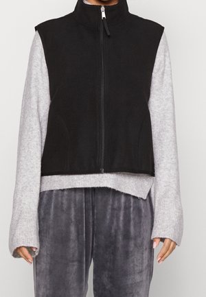 Veste noire sans manches zippée portée par-dessus un pull gris clair à manches longues et un pantalon en velours gris foncé sur une personne debout.