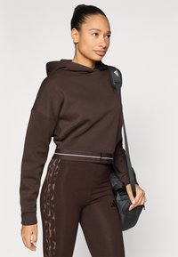 Καφέ κοντή κουκούλα με φαρδιά γιακά, σε συνδυασμό με ταιριαστά κολάν που έχουν πάνελ με λεοπάρ τύπωμα. Τσάντα κρεμασμένη πάνω σε έναν ώμο.