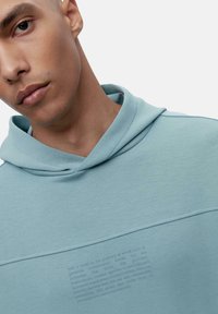 Hele sinine kapuutsiga särg, millel on tekstuuriga graafiline disain rinnal, varustatud reguleeritava kapuutsiga ja sileda kangaga. Kausis on peened õmblusdetailid.