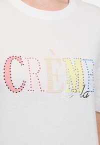 Valkoinen puuvillapaita, jossa on "CRÈME de la" monivärisillä rhinestone-kivillä. Tekstissä on vaaleanpunaisia, keltaisia, sinisiä ja mustia yksityiskohtia. Pyöreä kaula-aukko.