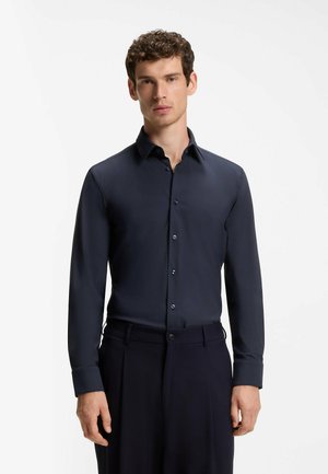 Uomo che indossa una camicia aderente blu navy scuro con bottoni e maniche lunghe e pantaloni neri su uno sfondo bianco semplice.