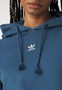 Zils kapucis, izgatavots no mīksta auduma, ar balto Adidas logo uz krūšu un aukliņām ar pomponiem. Tuvplāns uz kakla izgriezuma un auduma tekstūru.