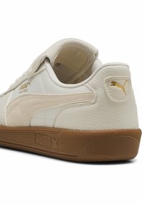 Bílé kožené tenisky s světle béžovými akcenty. Mají texturovanou hnědou podrážku, zlatý logo detail a zapínání na suchý zip u kotníku.