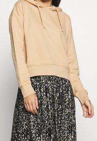 Beige cropped hoodie met trekkoorden, zachte textuur en geribbelde manchetten, gecombineerd met een zwarte plissérok met bloemenprint in tinten van room en bruin.