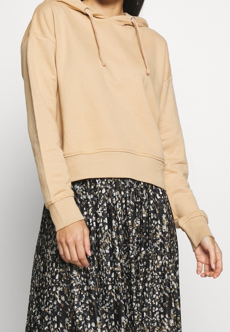 Beige cropped hoodie met trekkoorden, zachte textuur en geribbelde manchetten, gecombineerd met een zwarte plissérok met bloemenprint in tinten van room en bruin.