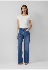 Haut court en côte blanc à manches courtes associé à un jean flare bleu clair taille haute. La mannequin se tient avec assurance, les mains dans les poches, mettant en valeur l'ajustement.