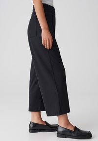 Pantalons larges noirs à taille haute, avec deux poches arrière et un tissu lisse et léger. Associés à des mocassins noirs.