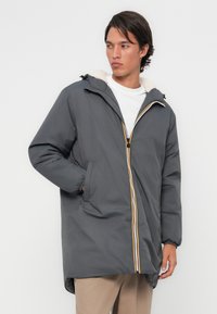 Manteau en duvet gris avec capuche, présentant une doublure blanche en peluche, une fermeture éclair à l'avant avec des accents jaunes et des poignets élastiqués.