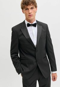 Noor mees musta satiinäärtega smokingus, valges triiksärgis ja musta kikilipsuga, seismas ühe käega pihus.