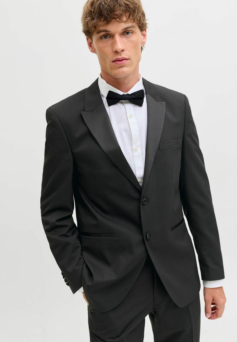 Noor mees musta satiinäärtega smokingus, valges triiksärgis ja musta kikilipsuga, seismas ühe käega pihus.